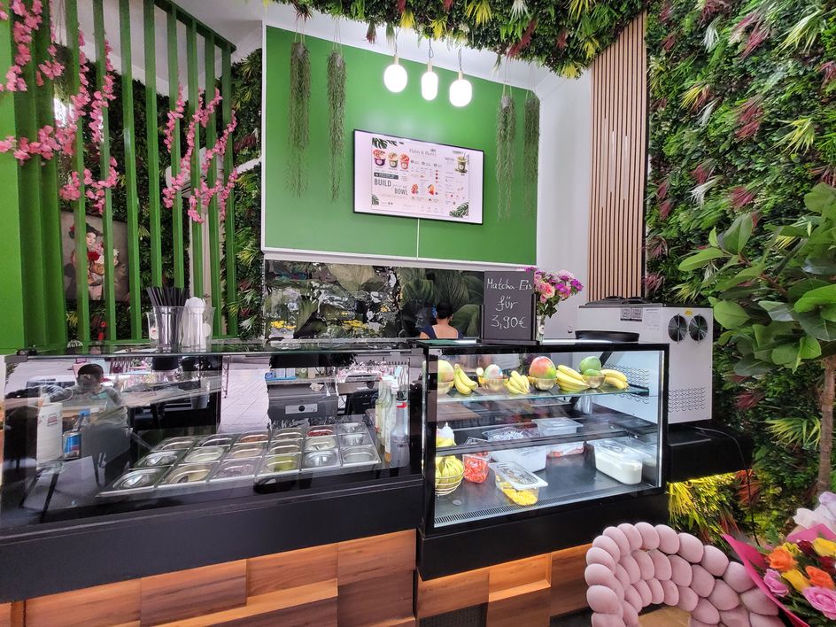 Blick in den Laden: Theke mit Toppings, Kühlvitrine voller frischem Obst und grüne Pflanzenwand